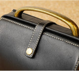 Close-up of a black leather handbag with white contrast stitching, a brass handle, and a matching brass snap strap.